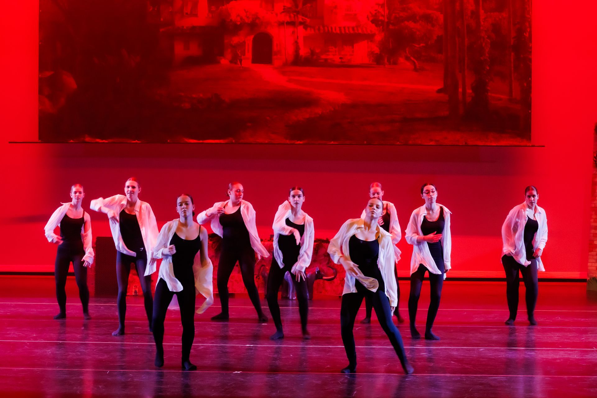 Dansers in zwarte maillots en witte shirts op een podium, verlicht met rode lampen.