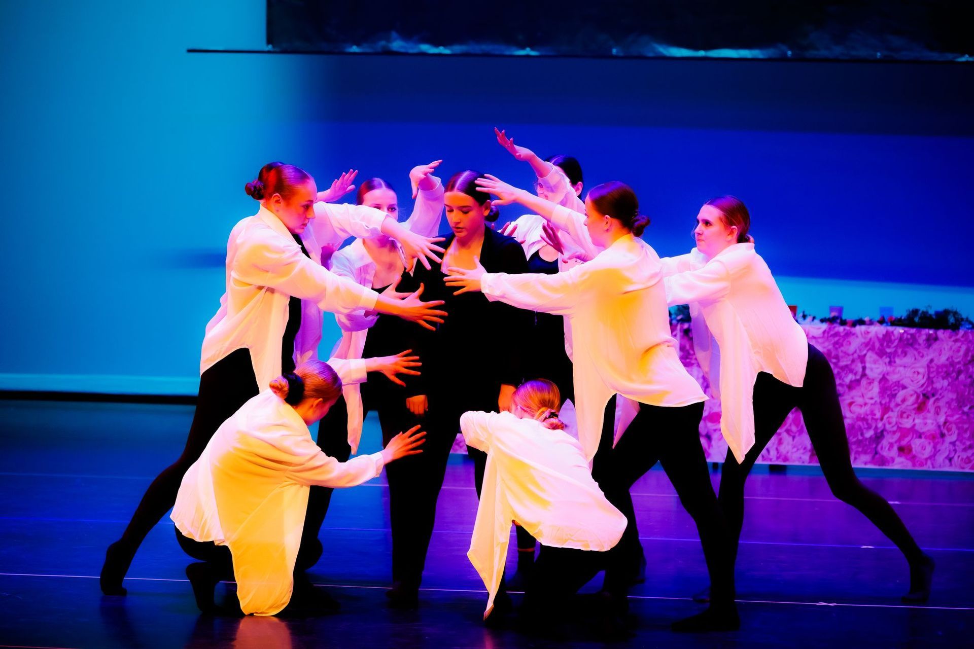 Dansers in witte shirts en zwarte broeken omringen een persoon in het zwart, met uitgestrekte handen. Blauwe podiumverlichting.
