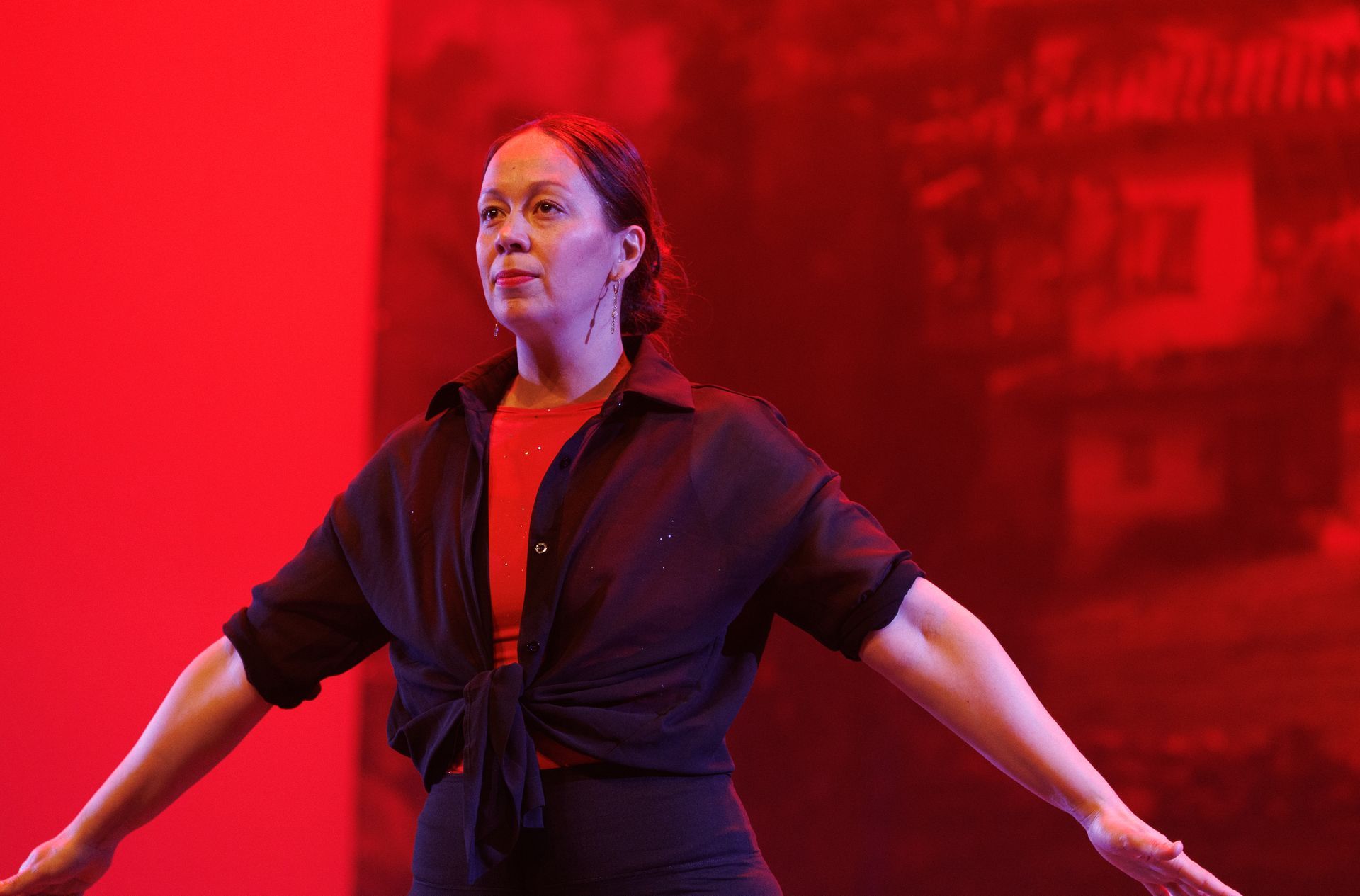 Vrouw in een zwart shirt en een rode top, met uitgestrekte armen, op een rood podium met een wazige achtergrond.