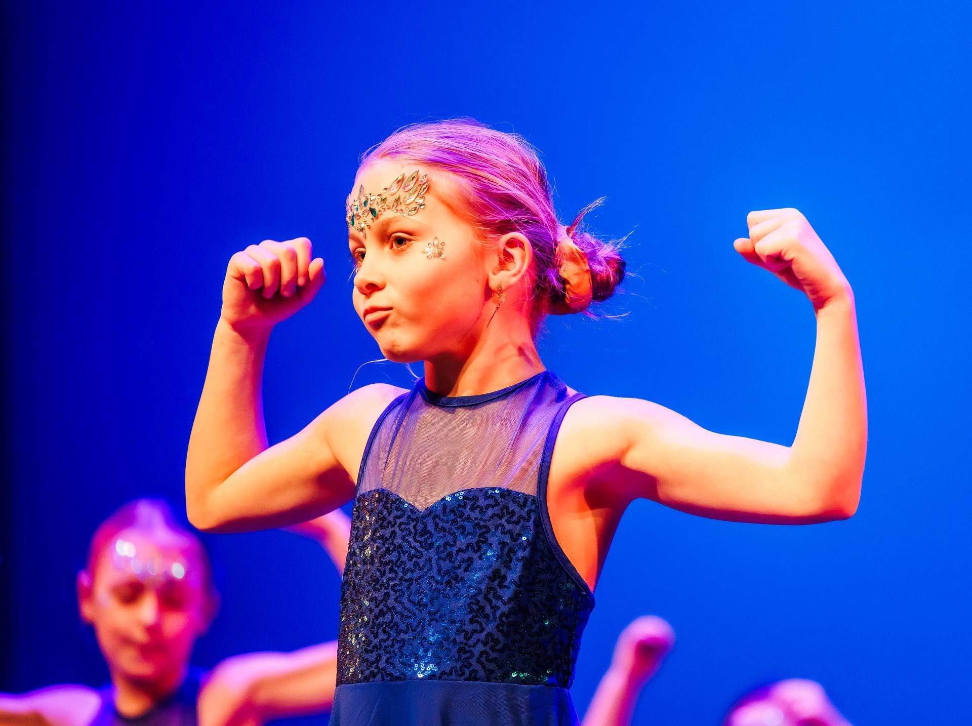 Meisje in een blauw danskostuum spant haar armen op het podium bij blauwe verlichting.