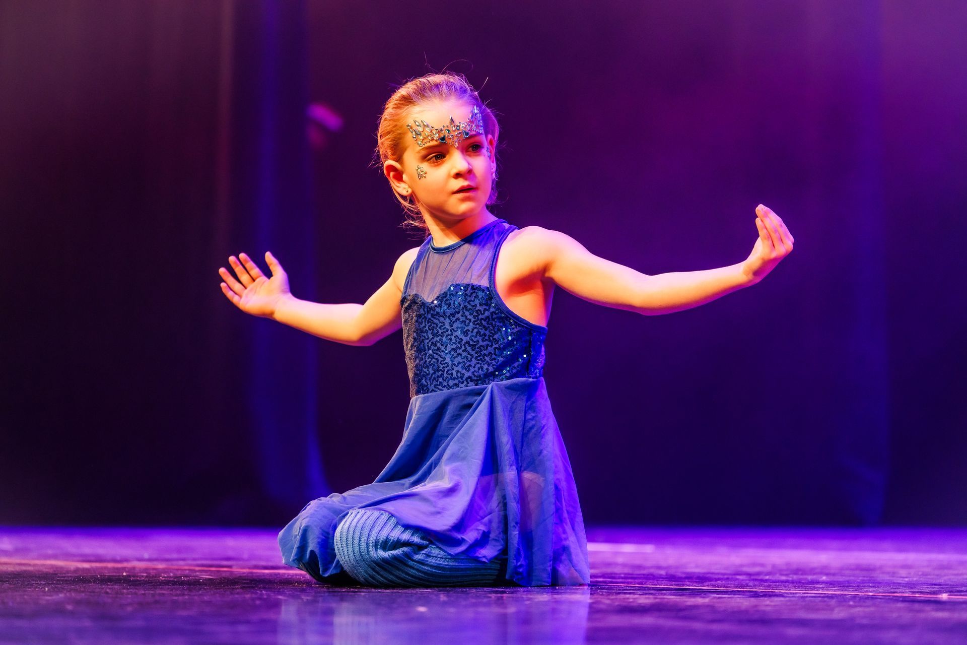 Jonge danseres in een blauwe jurk knielt op het podium met uitgestrekte armen.