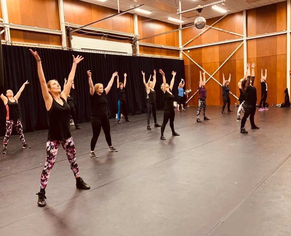 Een groep mensen in een dansles met de armen omhoog. In een grote zaal.