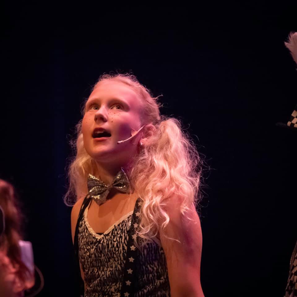 Meisje met blonde vlechtjes en een vlinderdasje, zingend op het podium, in de spotlights, tegen een donkere achtergrond.
