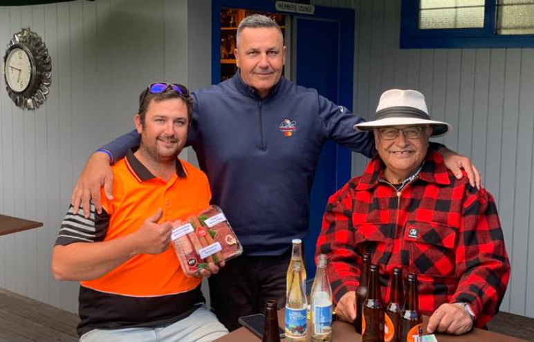 Three men are posing for a picture while sitting at a table with bottles of beer.