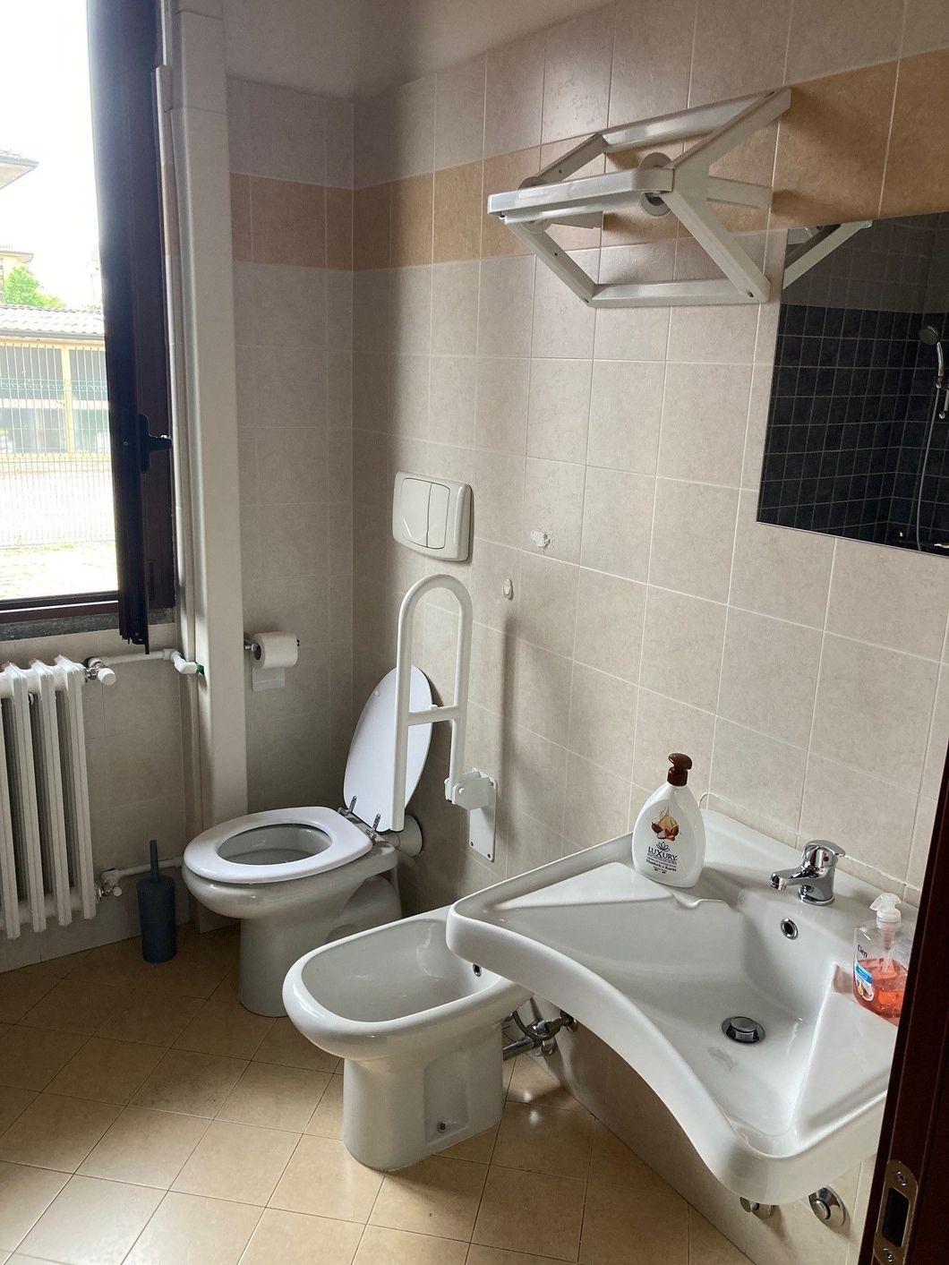 Bagno con WC, bidet, lavandino e finestra, con corrimano accessibile. Pareti piastrellate beige.