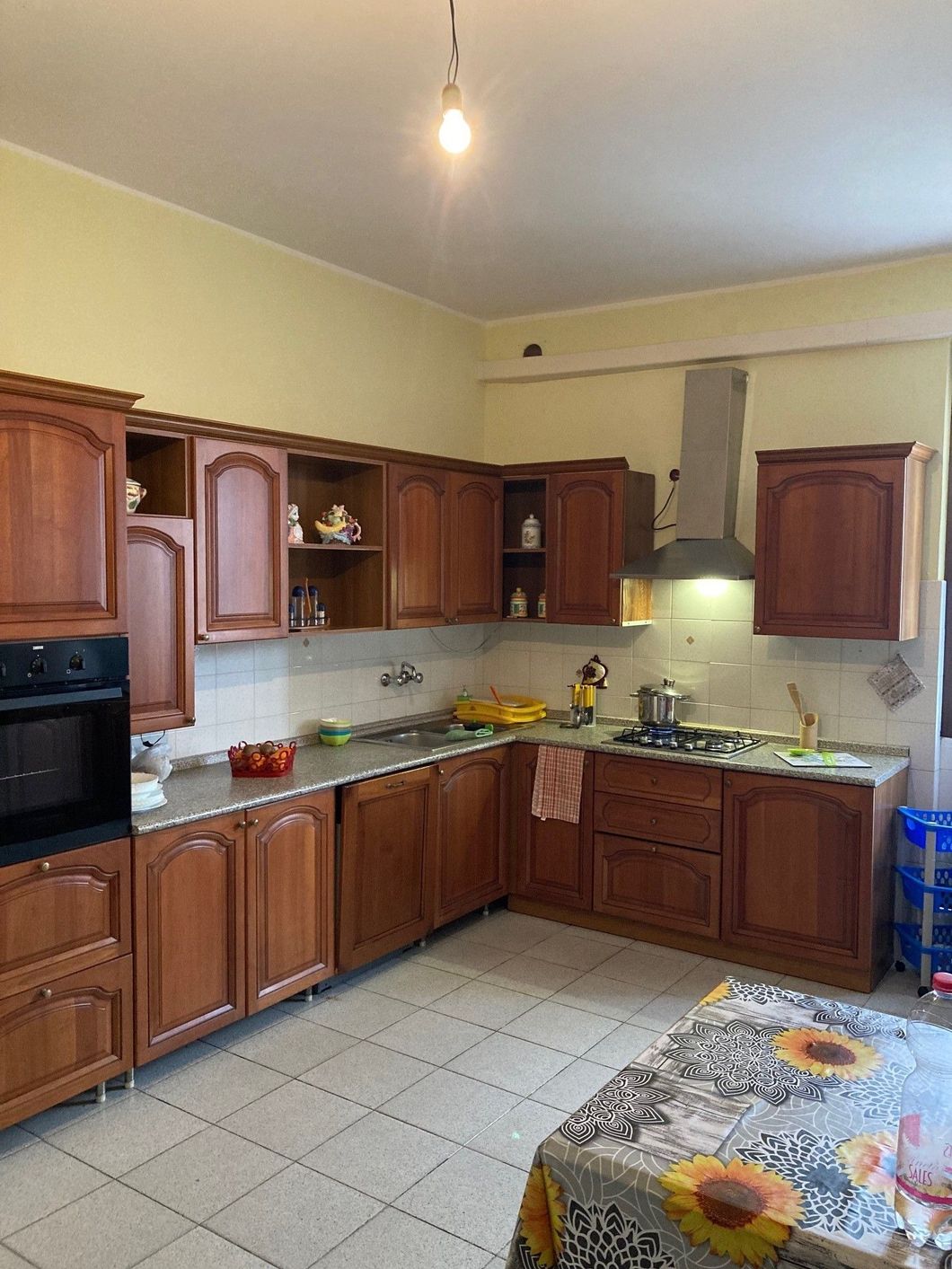 Cucina con mobili marroni, pareti e ripiani chiari e un tavolo con una tovaglia a fiori.
