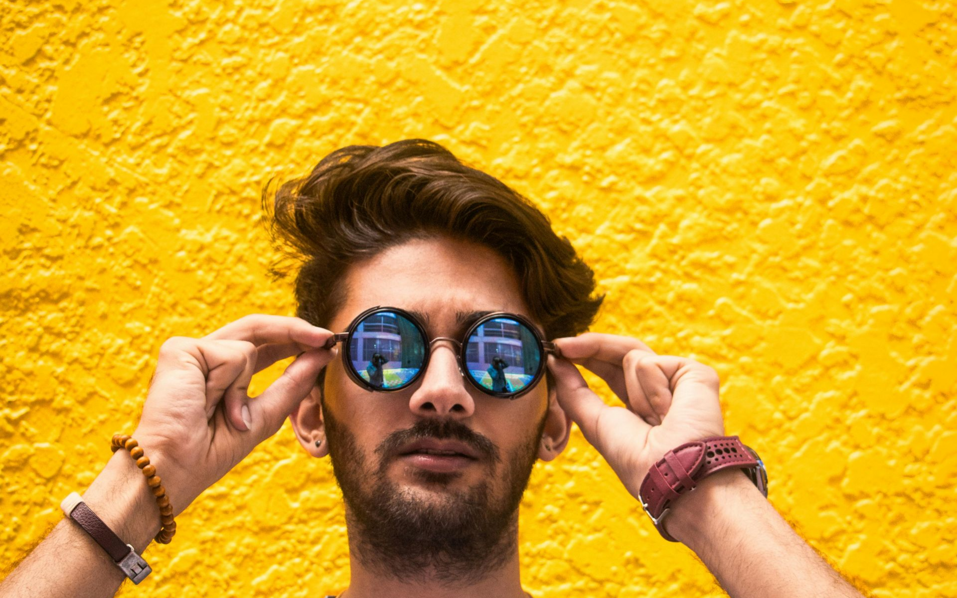 Man in round sunglasses against a yellow wall, adjusting the frames.