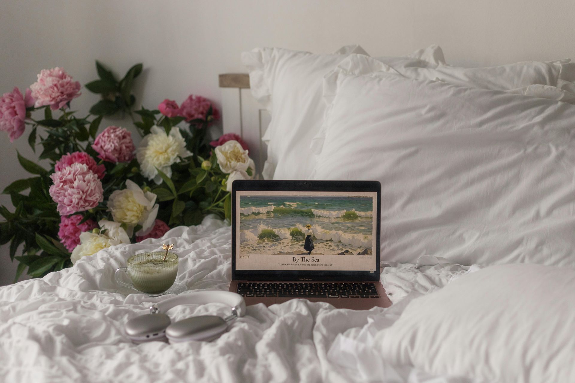 Laptop on bed with flowers, drink, and headphones. The screen shows a beach scene, all in a cozy, bright setting.
