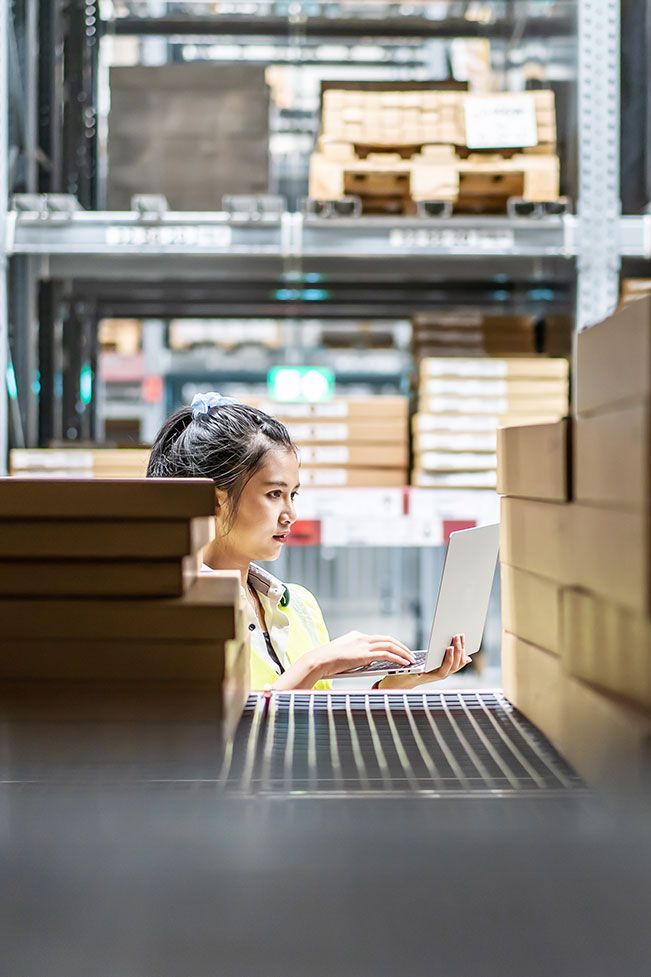 Person working on a laptop in a warehouse aisle surrounded by stacked boxes and pallets