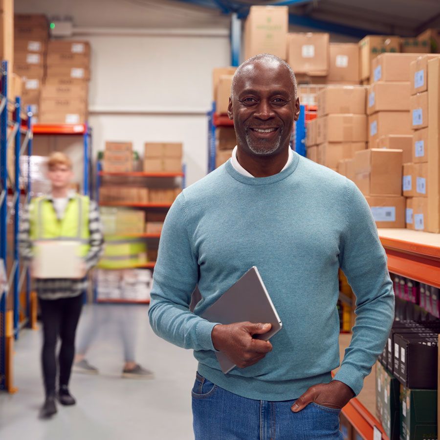 A portrait of a man holding a digital laptop in a busy warehouse. There are shelves of boxes and employees behind him.