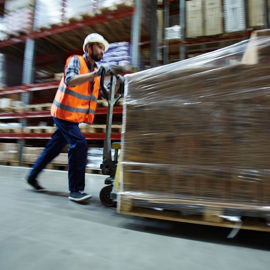 Warehouse worker in an orange vest pushing a pallet jack with wrapped boxes on a pallet