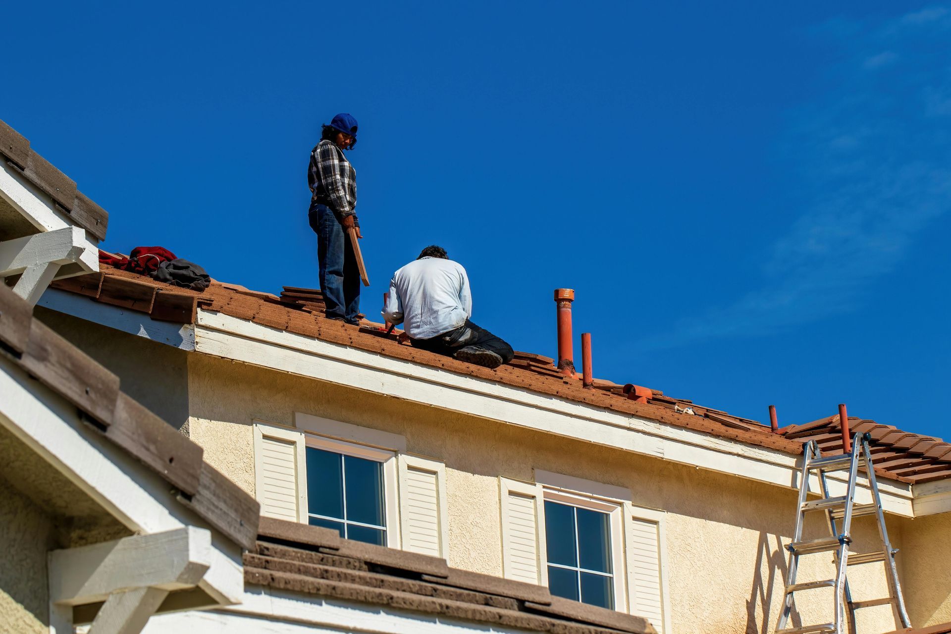 Two workers on a rooftop repairing tiles under a clear blue sky