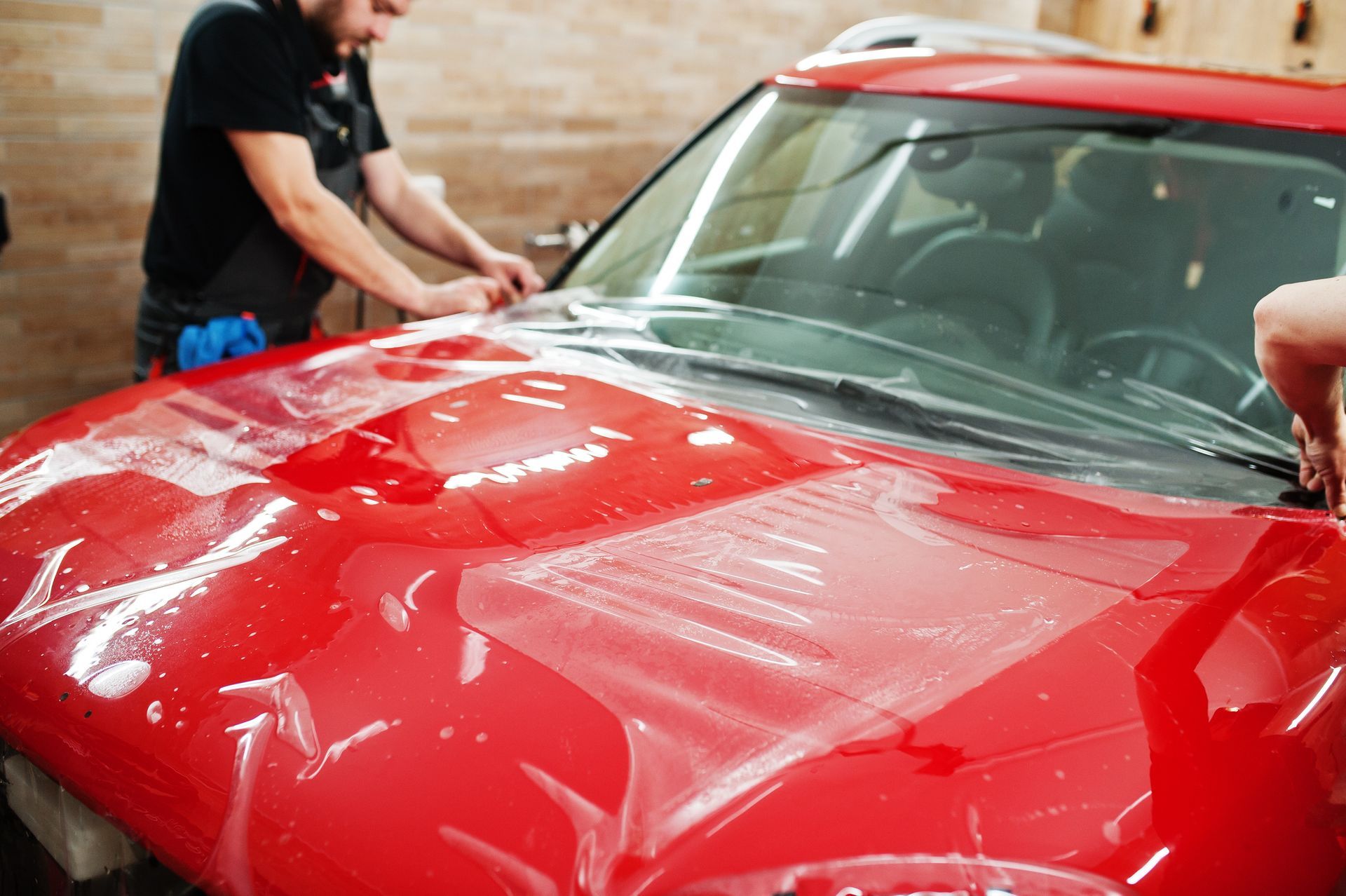 A man is applying a protective film to the hood of a red car.