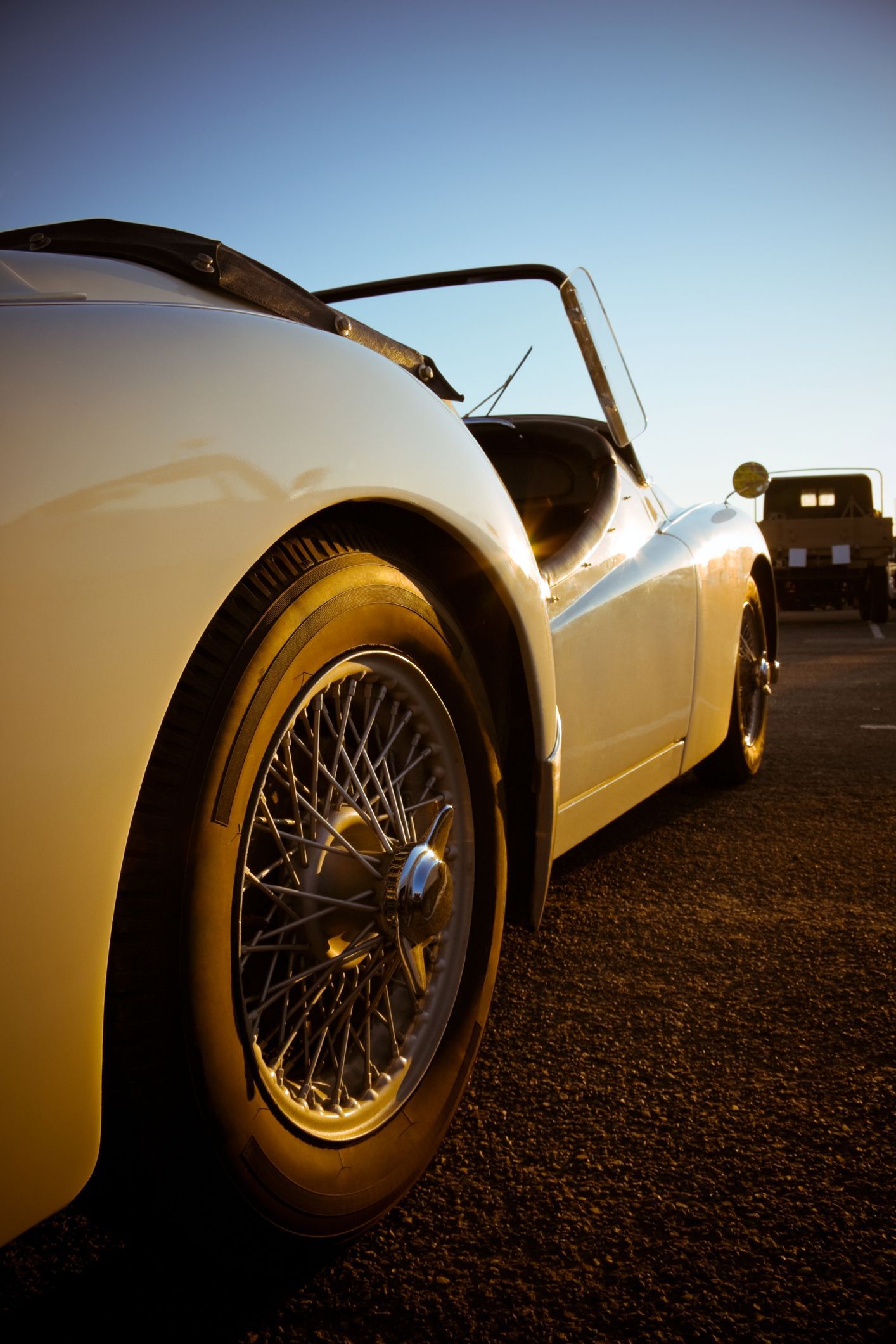 Cream-colored classic convertible car parked on asphalt, wire wheels, blue sky backdrop.