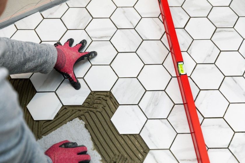 Hands in red gloves installing white hexagonal floor tiles beside a red level tool.