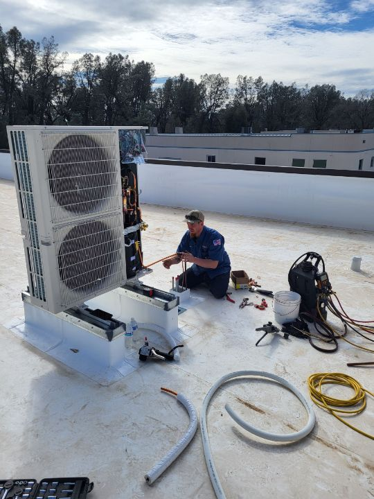 A man is kneeling on a roof working on an air conditioner.