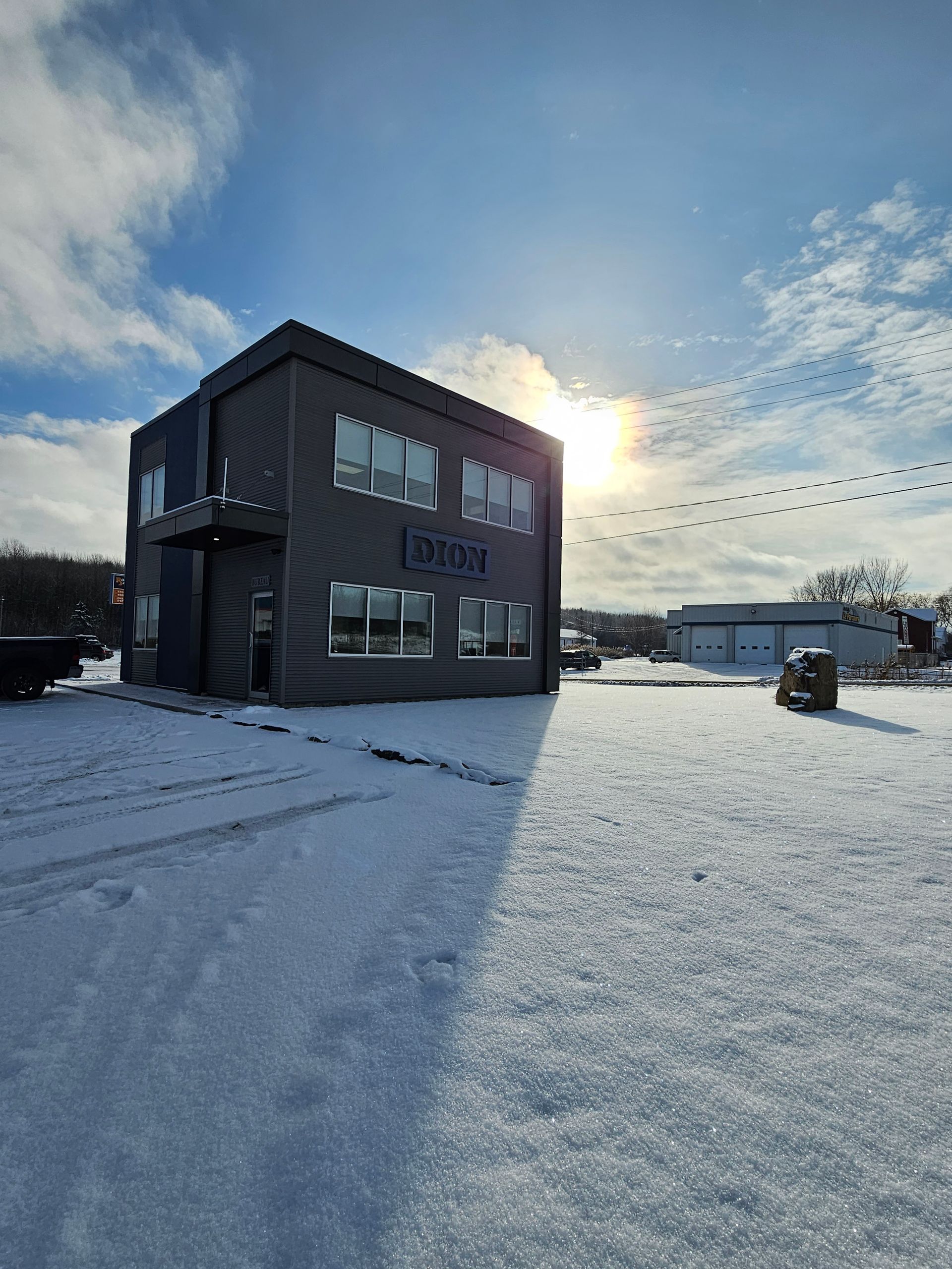 Champ enneigé à côté d'un bâtiment sombre au lever du soleil, avec des empreintes de pas dans la neige et un ciel d'hiver lumineux.