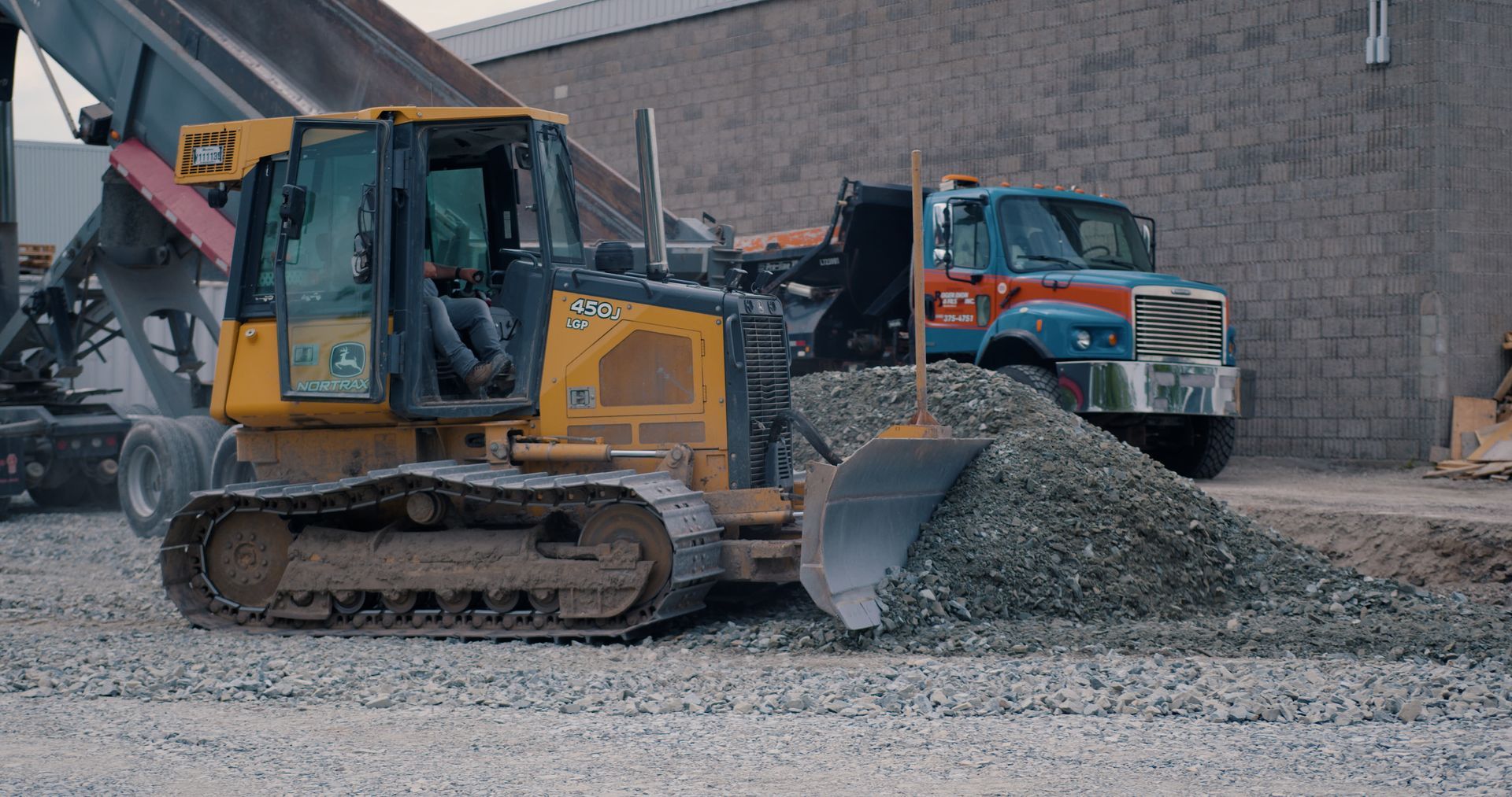 Une pelleteuse jaune à côté d'un tas de gravier sur un chantier, avec un camion-benne orange en arrière-plan.