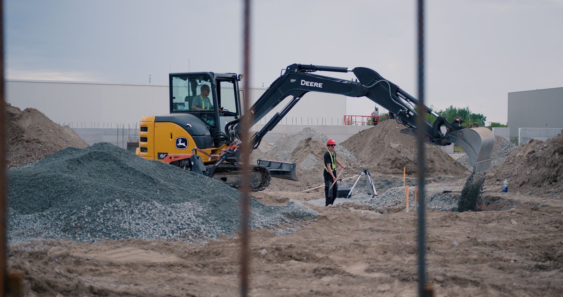 Une excavatrice travaille sur un site d'extraction de gravier, un ouvrier se tient à proximité et des tas de terre sont visibles en arrière-plan.