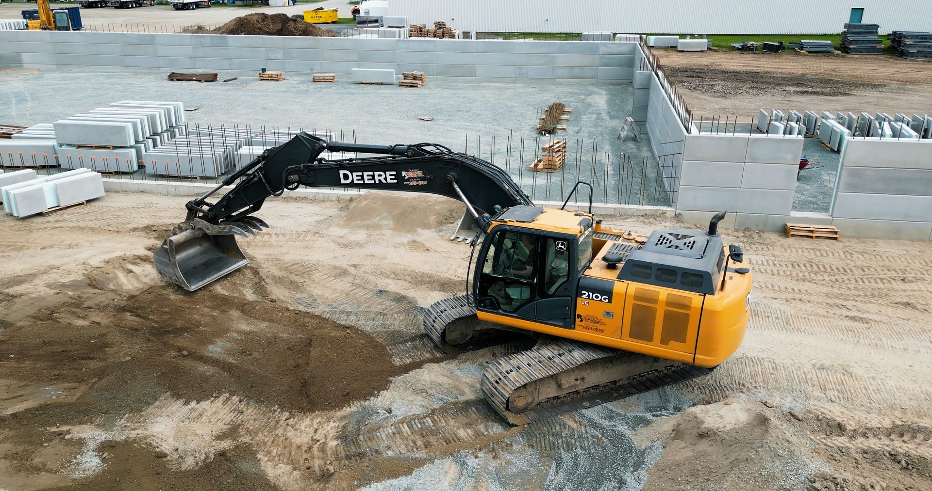 Une pelleteuse jaune déplace de la terre sur un chantier de construction avec des murs en béton et du matériel en arrière-plan.