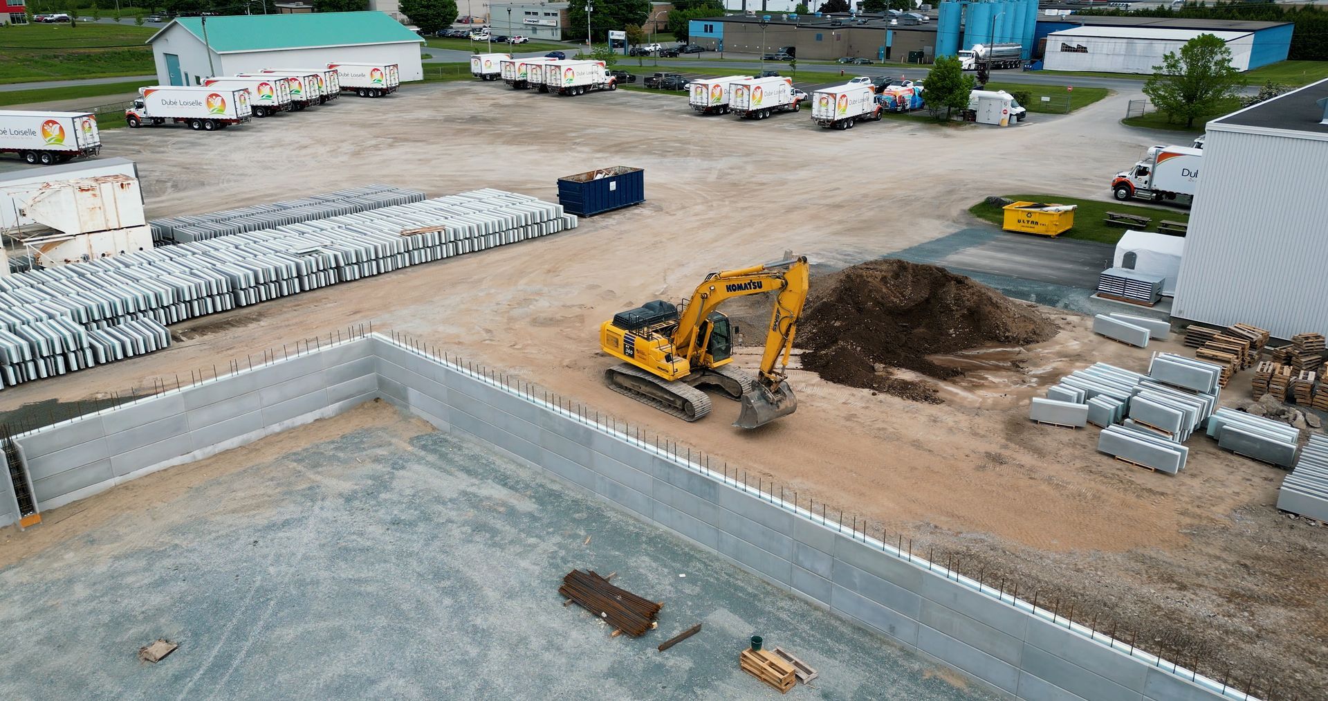 Chantier de construction avec une pelle mécanique jaune creusant près de fondations en béton et de piles de matériaux