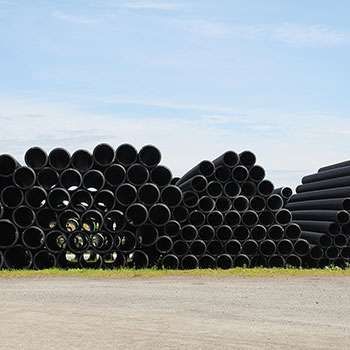 Des piles de tuyaux noirs alignés à l'extérieur, sous un ciel dégagé, à côté d'une zone pavée.