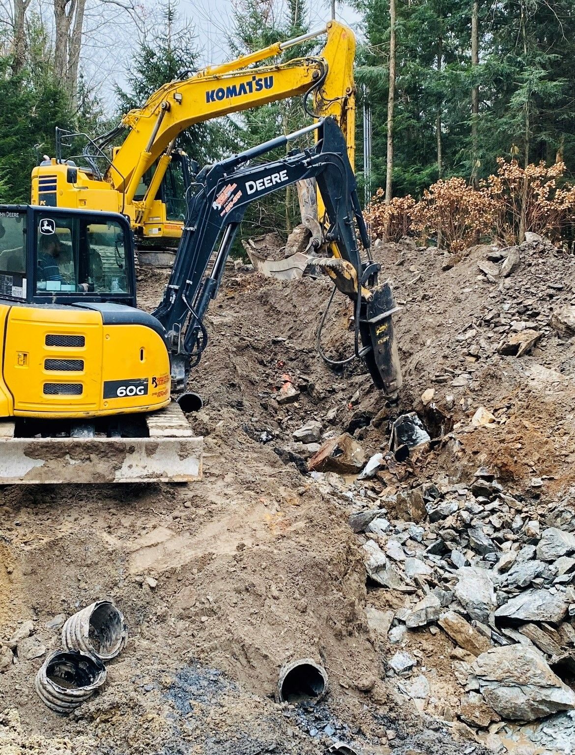 Une pelleteuse jaune creuse dans une tranchée de construction boueuse, avec des tuyaux et des rochers apparents, entourée d'arbres.