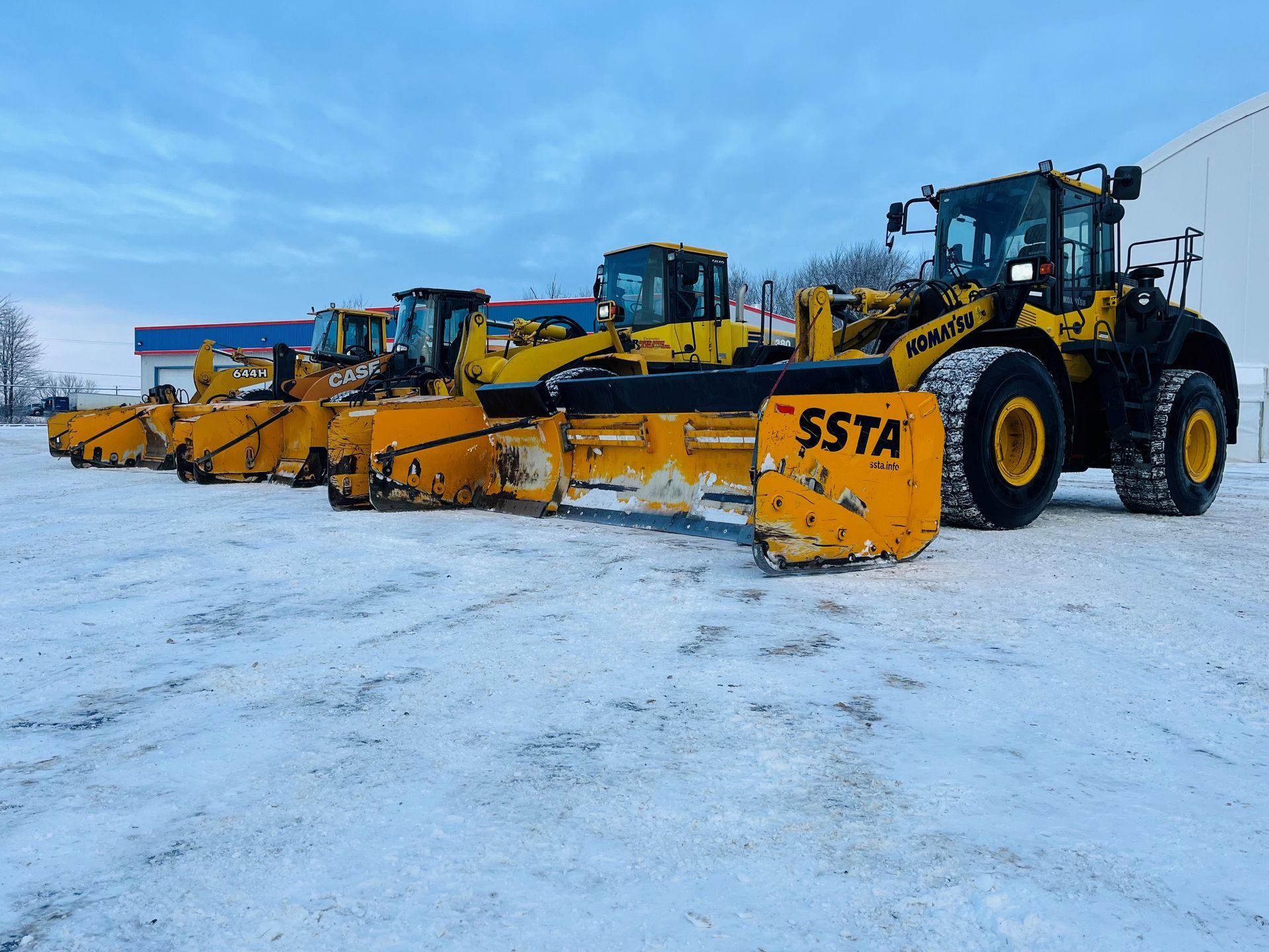 Une rangée de chasse-neige et de chargeuses frontales jaunes stationnées sur un terrain enneigé sous un ciel bleu