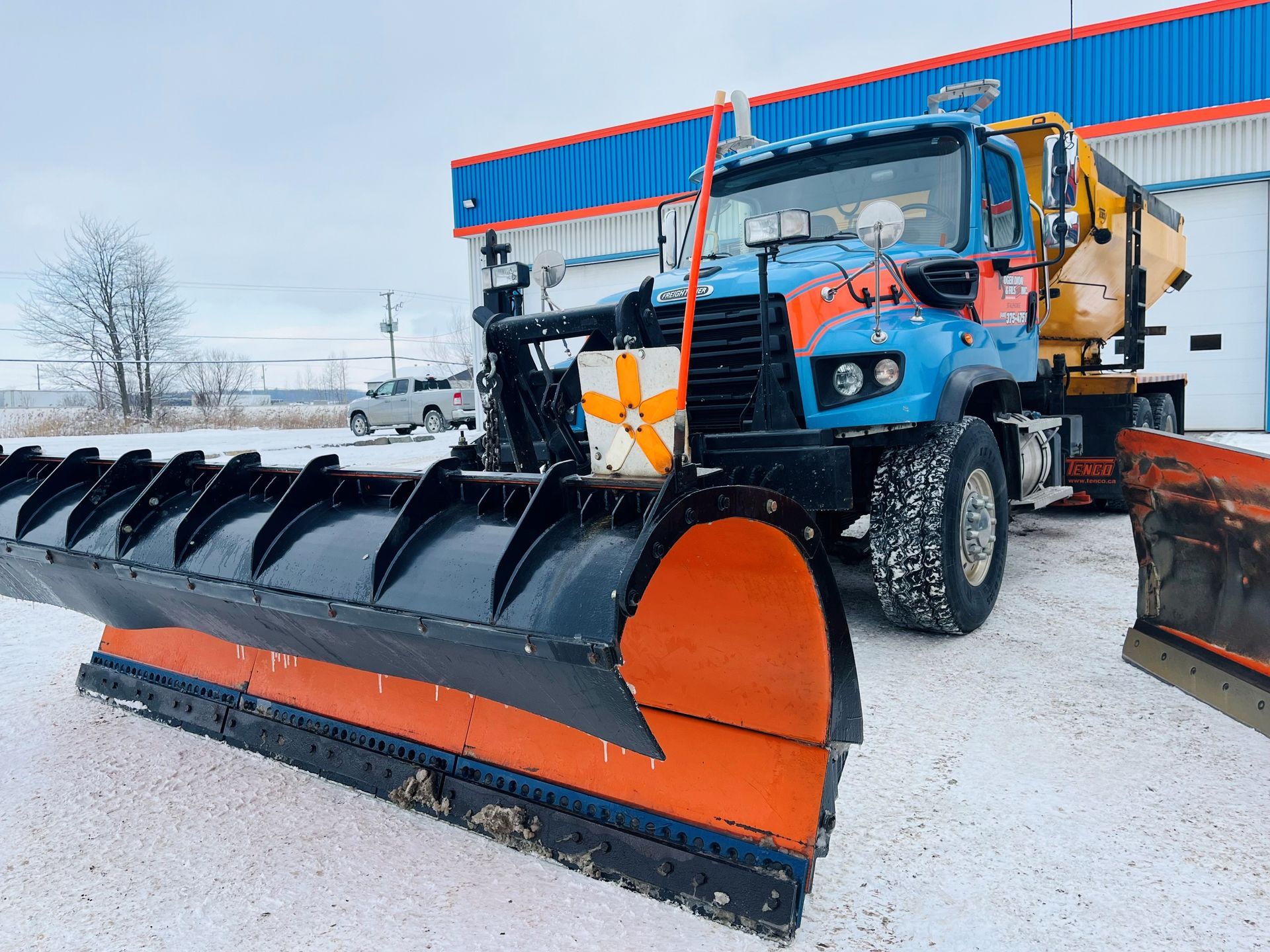 Un camion chasse-neige bleu avec une lame orange est stationné sur un terrain enneigé.