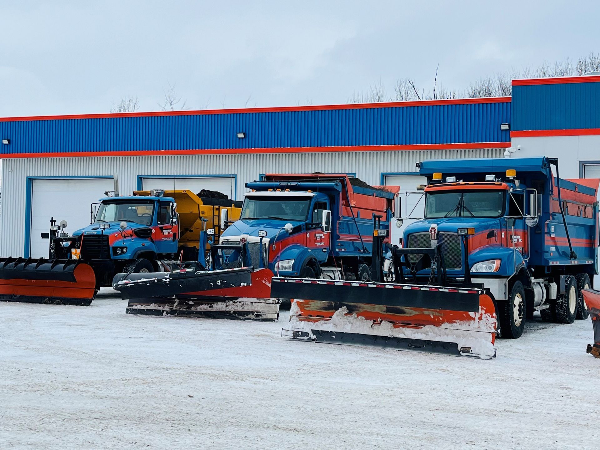 Des chasse-neige sont stationnés sur un terrain adjacent à un bâtiment industriel bleu.