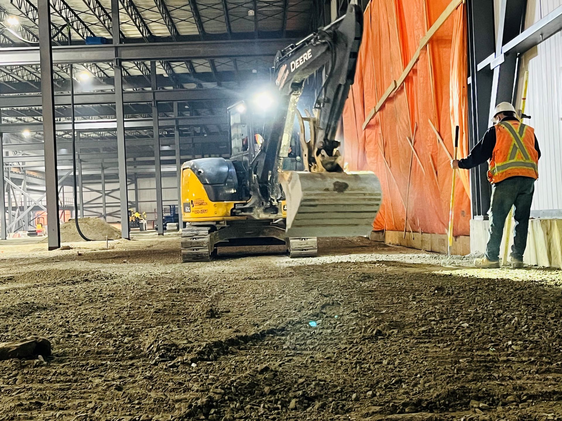Une excavatrice et un ouvrier à l'intérieur d'un chantier de construction d'entrepôt, délimité par des barrières de sécurité orange.