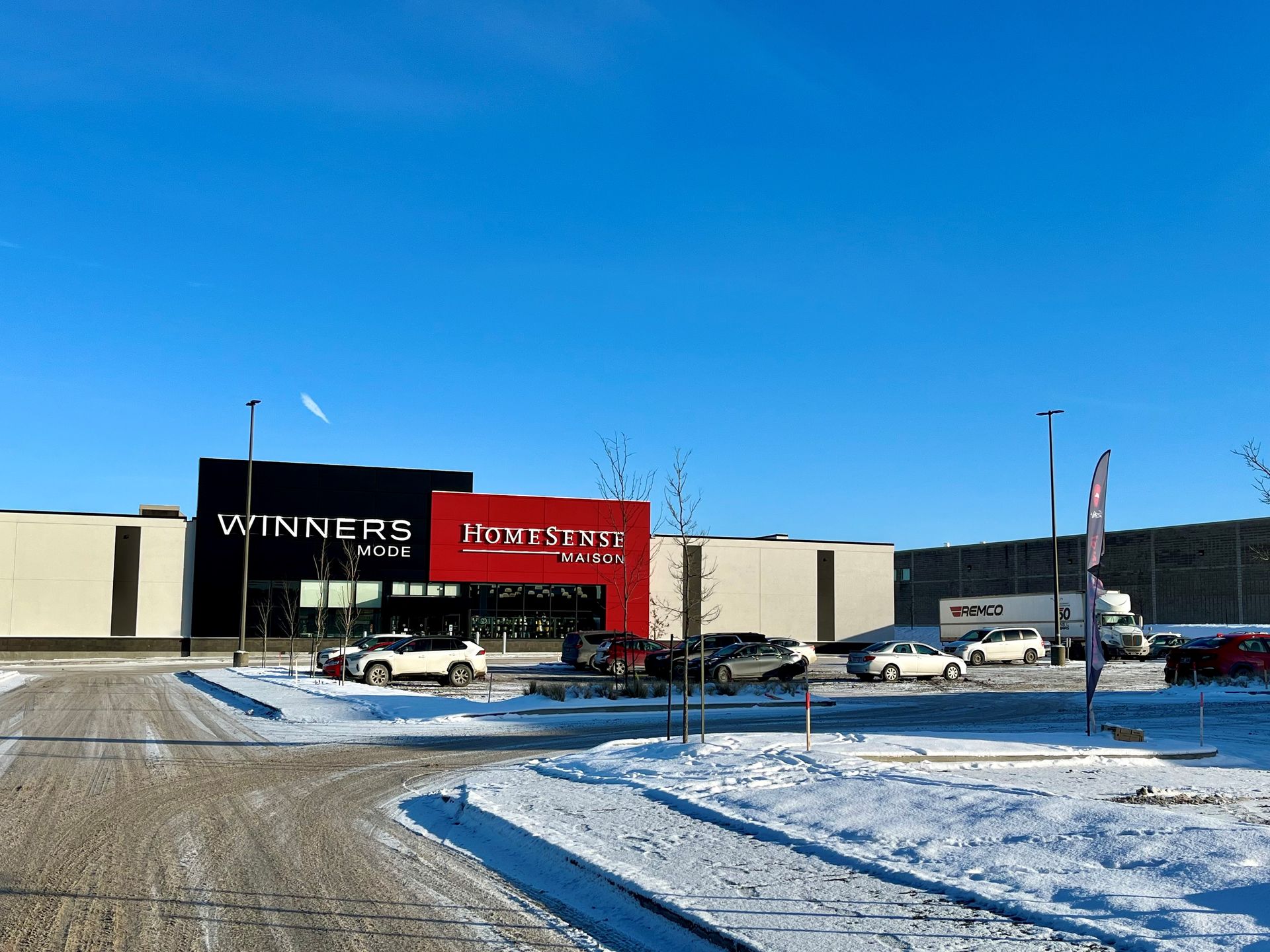 Place commerciale enneigée avec une façade rouge et des voitures garées sous un ciel bleu clair