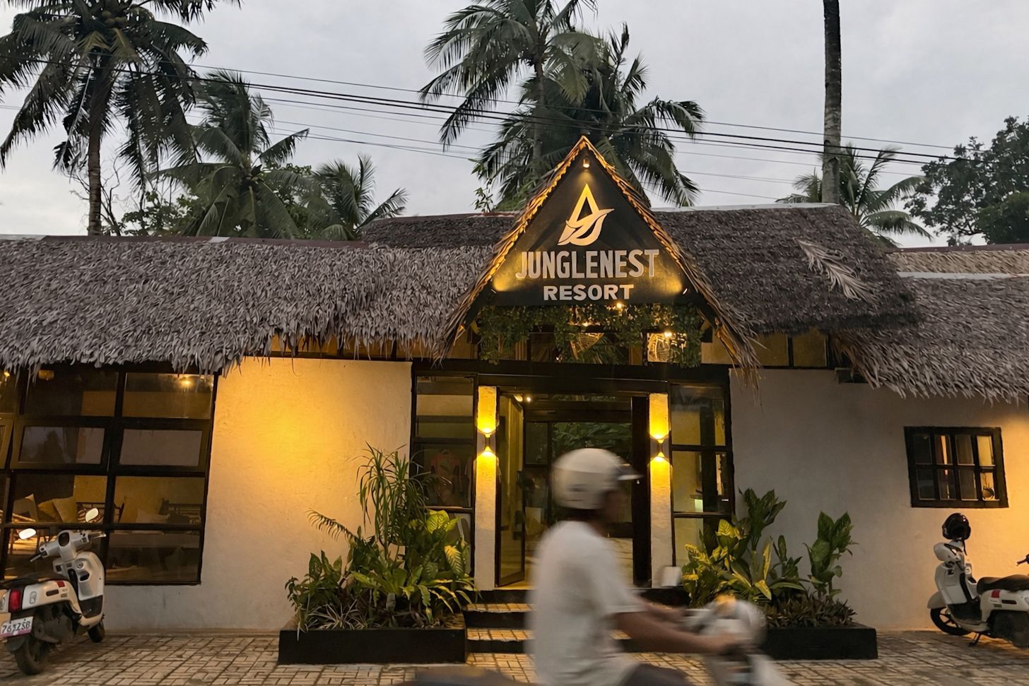 Thatched-roof restaurant at dusk with a lit sign and a blurred scooter passing in front