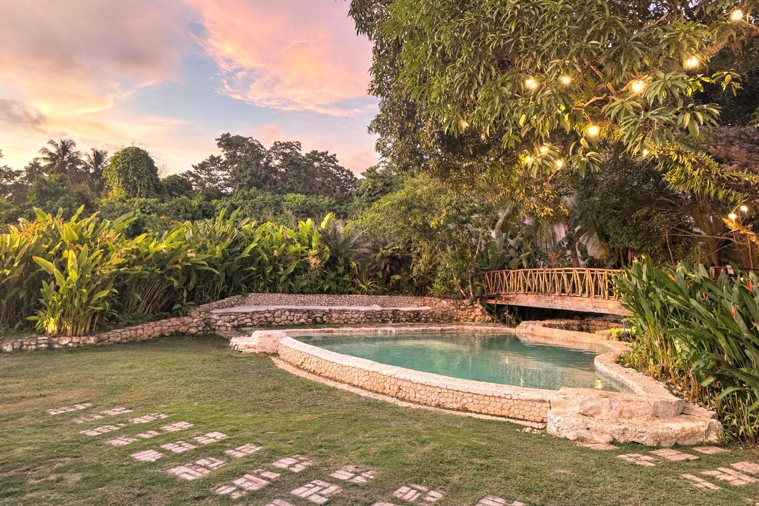 Stone-edged backyard pool beside a lit tree and wooden bridge at sunset