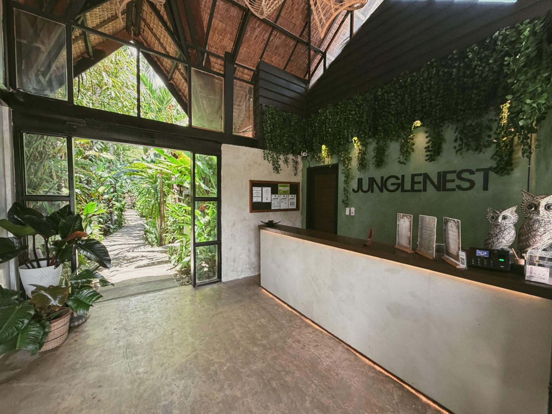 Modern lobby with a white reception desk, green wall logo, and open view to lush tropical greenery
