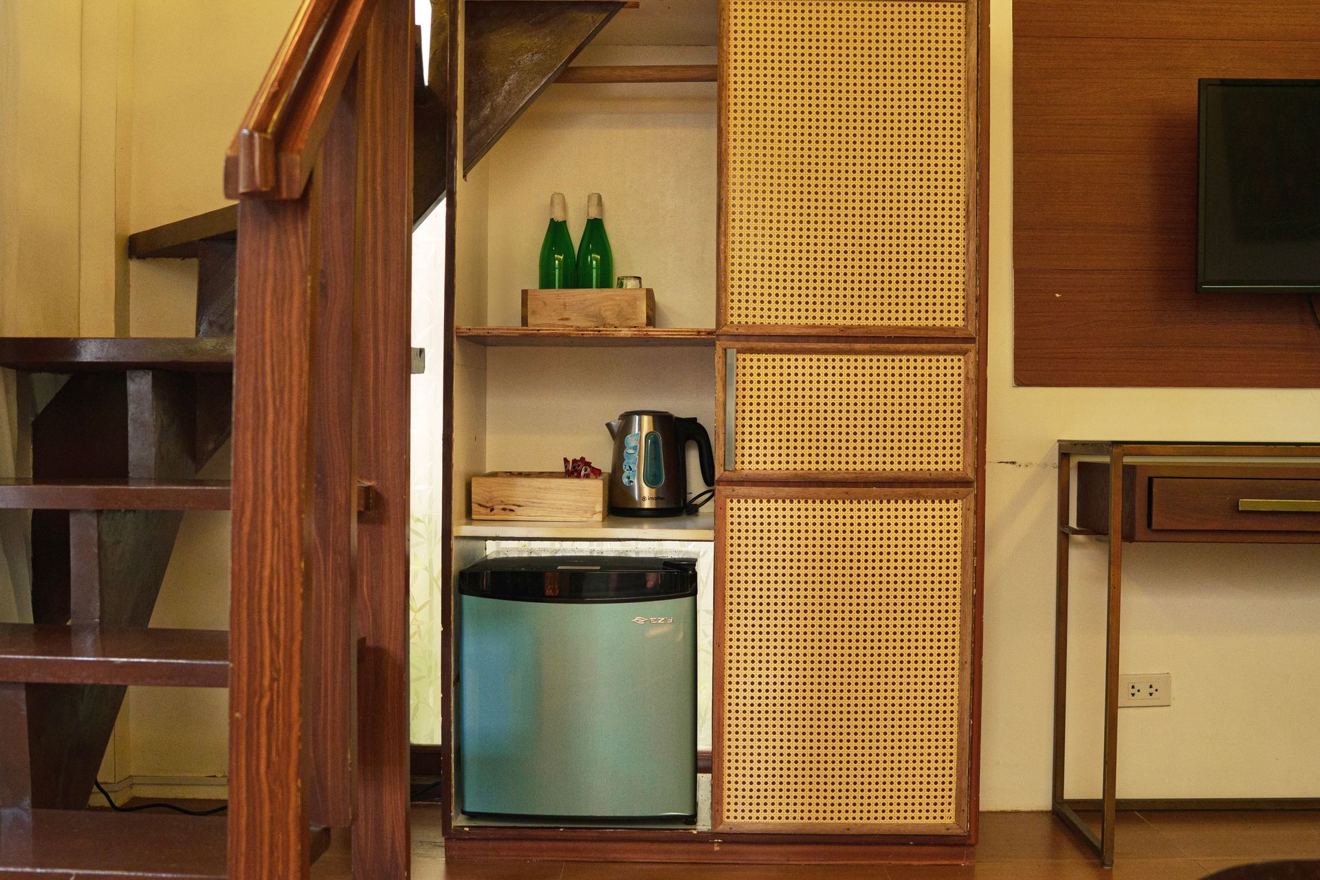 Small kitchenette with mini fridge, kettle, shelves, and woven cabinet doors in a room corner