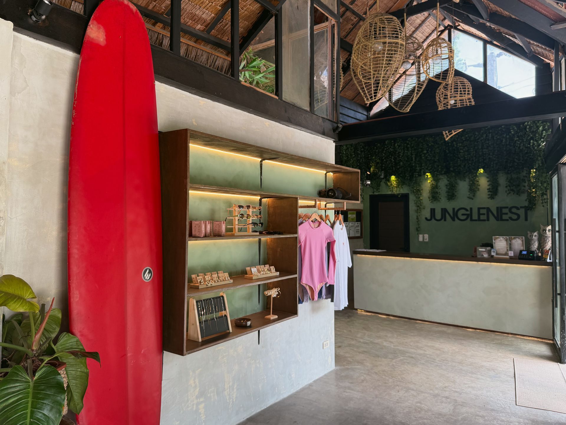 Surf shop interior with a red surfboard, display shelves, hanging clothes, and a reception counter.