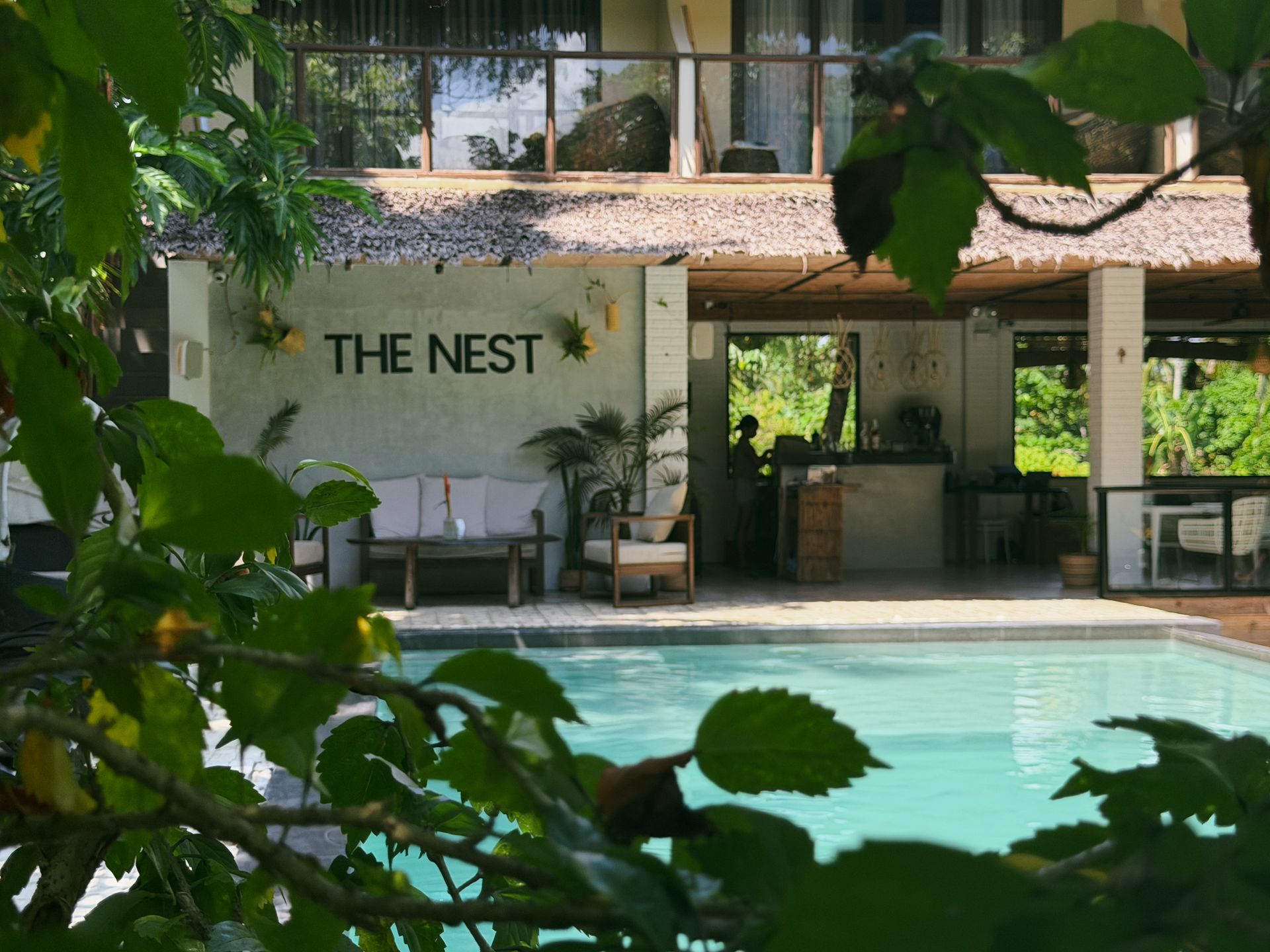 A tropical poolside café with the sign “THE NEST,” framed by lush green leaves.