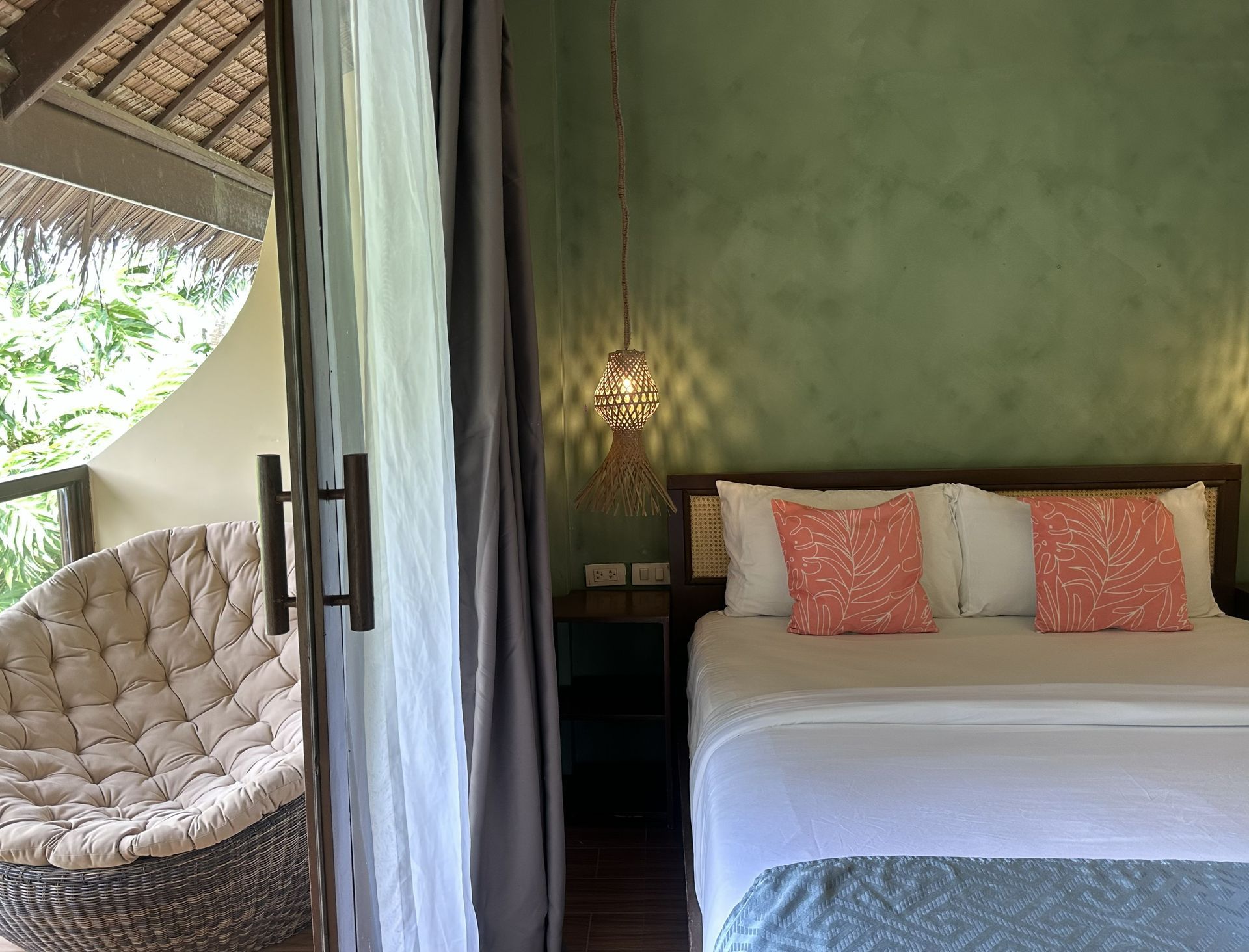 Tropical bedroom with white bed, coral pillows, green wall, and hanging chair by the sliding curtain.