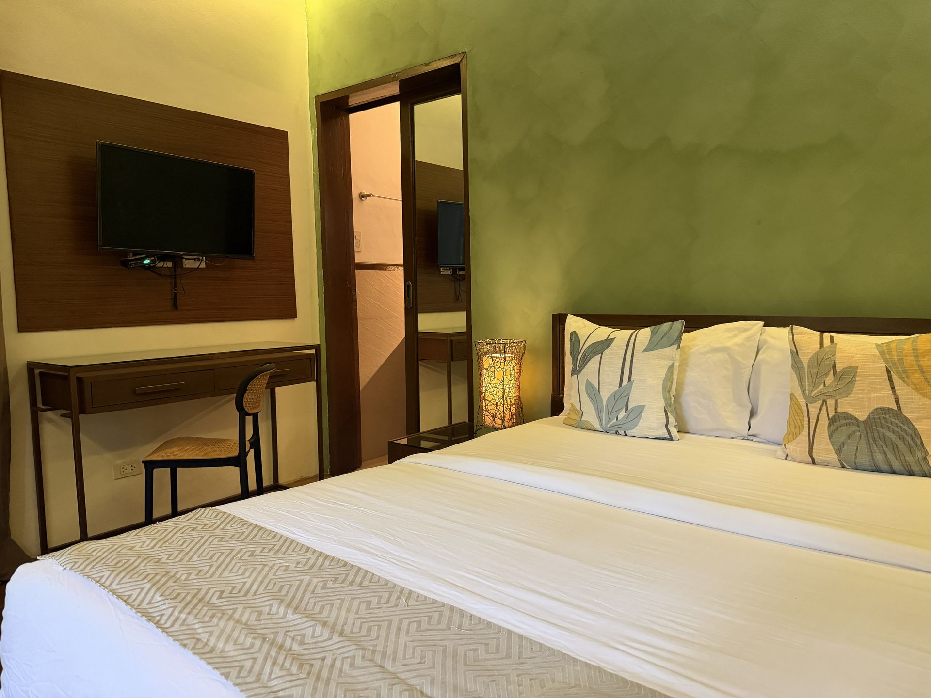Hotel room with a neatly made bed, wall-mounted TV, desk, and soft green walls.