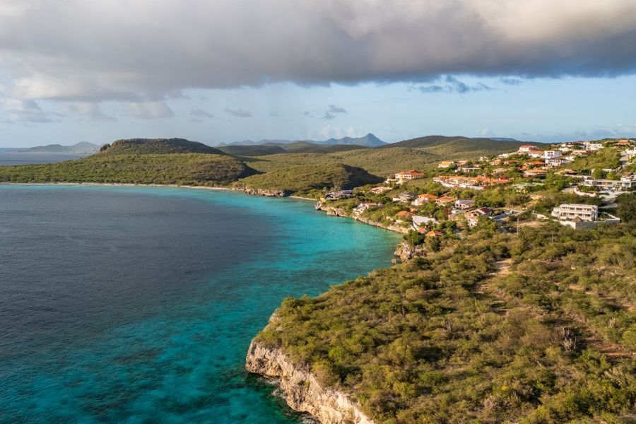 Luchtfoto van turquoise water dat een rotsachtige kust ontmoet, met huizen verscholen in het groen onder een bewolkte hemel.