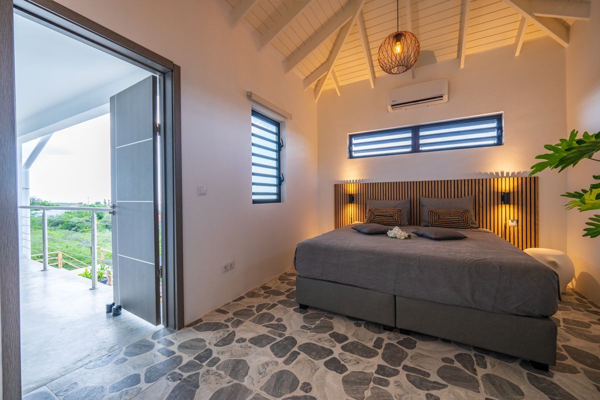 Slaapkamer met openslaande deur naar een balkon. Grijs bed, stenen vloer, witte muren en uitzicht op groen.