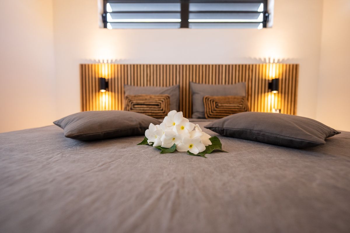 Slaapkamer met grijs beddengoed, houten hoofdbord, twee kussens, twee lampen, bloemen en een raam.