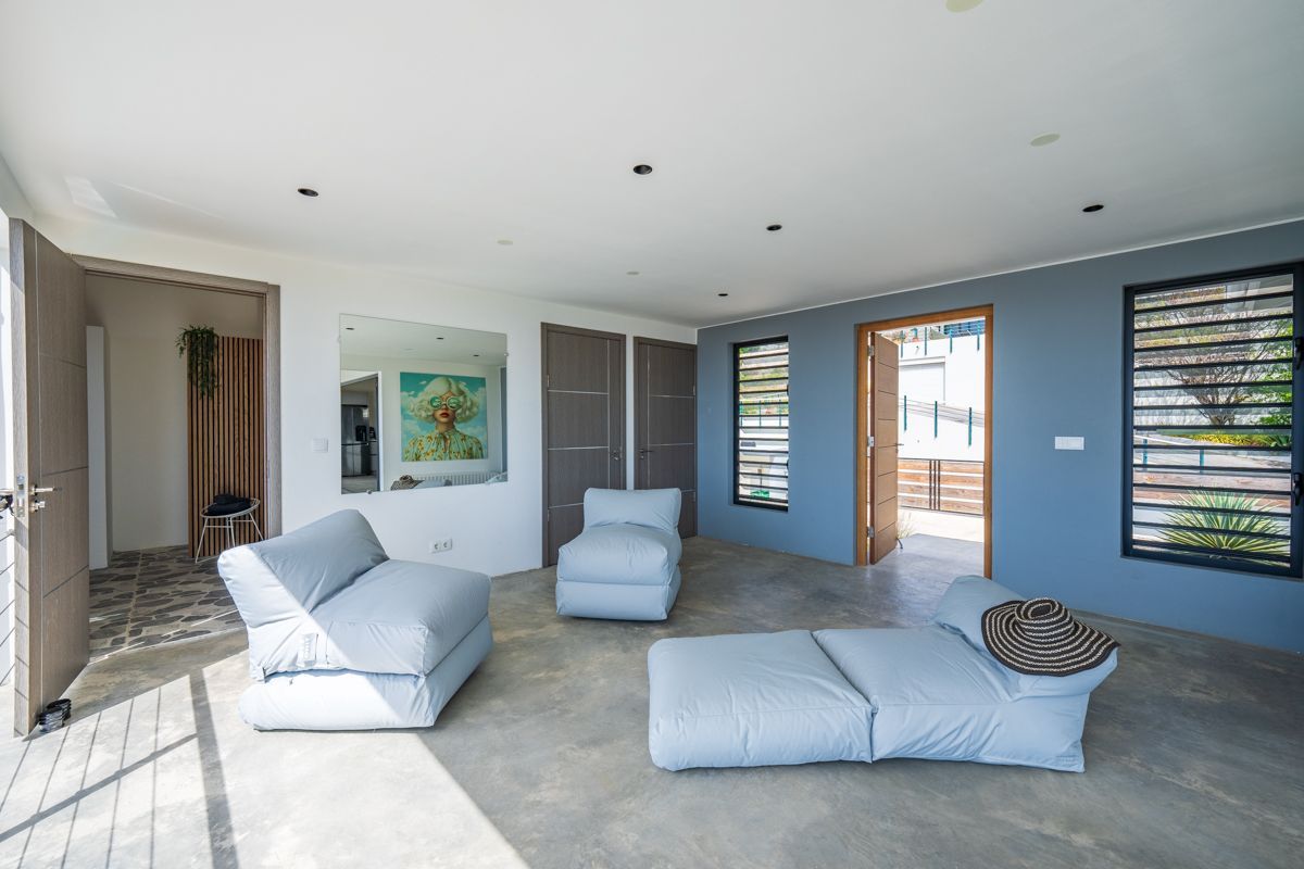 Zonnige kamer met blauwe muren, betonnen vloer en lichtblauwe loungebanken. Open deuren/ramen.