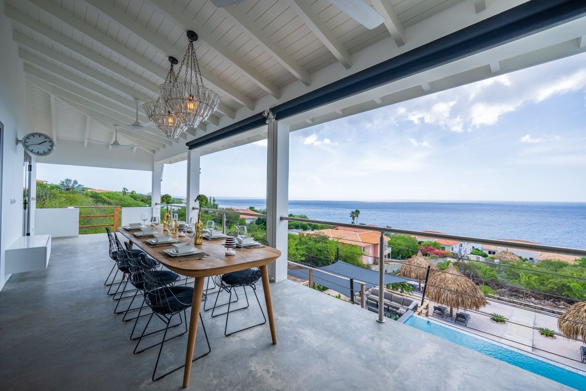 Overdekt terras met uitzicht op de oceaan, een lange tafel gedekt voor een maaltijd en een kroonluchter.