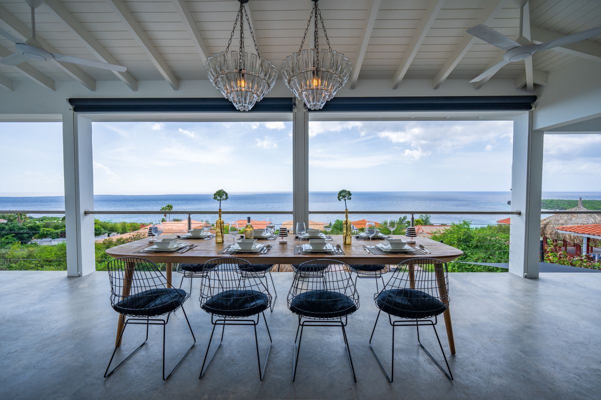 Eettafel op een terras met uitzicht op de oceaan; moderne stoelen, kristallen kroonluchters, blauwe lucht.