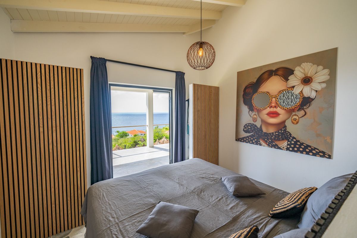 Slaapkamer met bed, kunst, raam met uitzicht op zee en houten accenten.