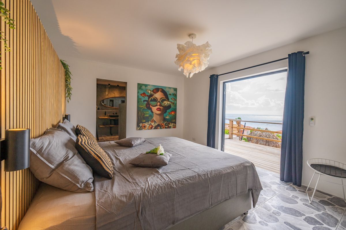 Slaapkamer met kingsize bed en openslaande deur naar het terras. Kunst aan de muur, grijs beddengoed. Plafondlamp en houten accentmuur.