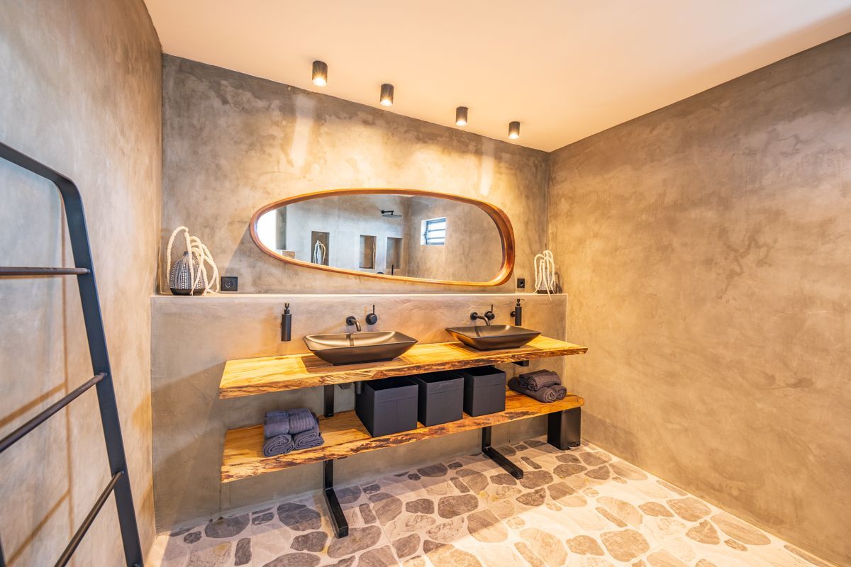 Moderne badkamer met een houten badmeubel, zwarte wastafels en een grote ovale spiegel. Grijze stenen vloer en wanden.