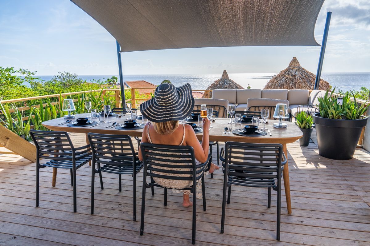 Vrouw met grote hoed zit aan een eettafel in de buitenlucht met uitzicht op de oceaan.